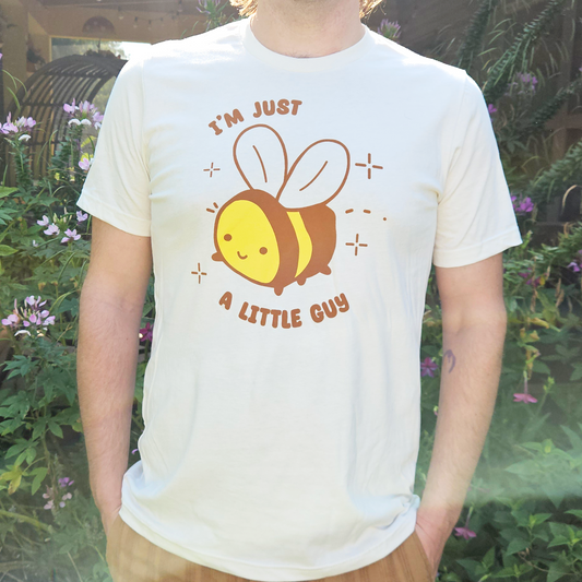 A man wearing a white graphic t-shirt with a cartoon bee and the text 'I'M JUST A LITTLE GUY' in brown, standing outdoors in front of a garden background.